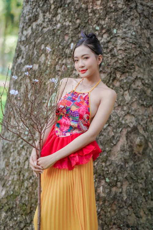 A woman in a yellow and red dress posing near a tree