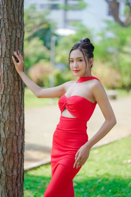 Asian woman posing elegantly in a red dress outdoors by a tree.