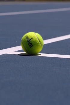 Bright green tennis ball resting on a blue court line under sunlight.