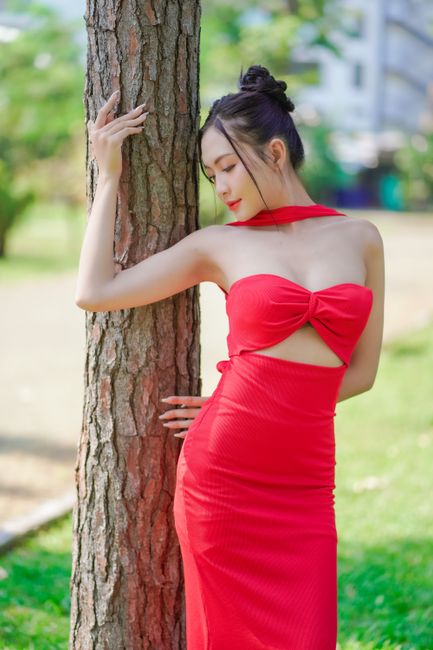 Stylish portrait of a young woman posing in a red dress against a tree in a summer park.