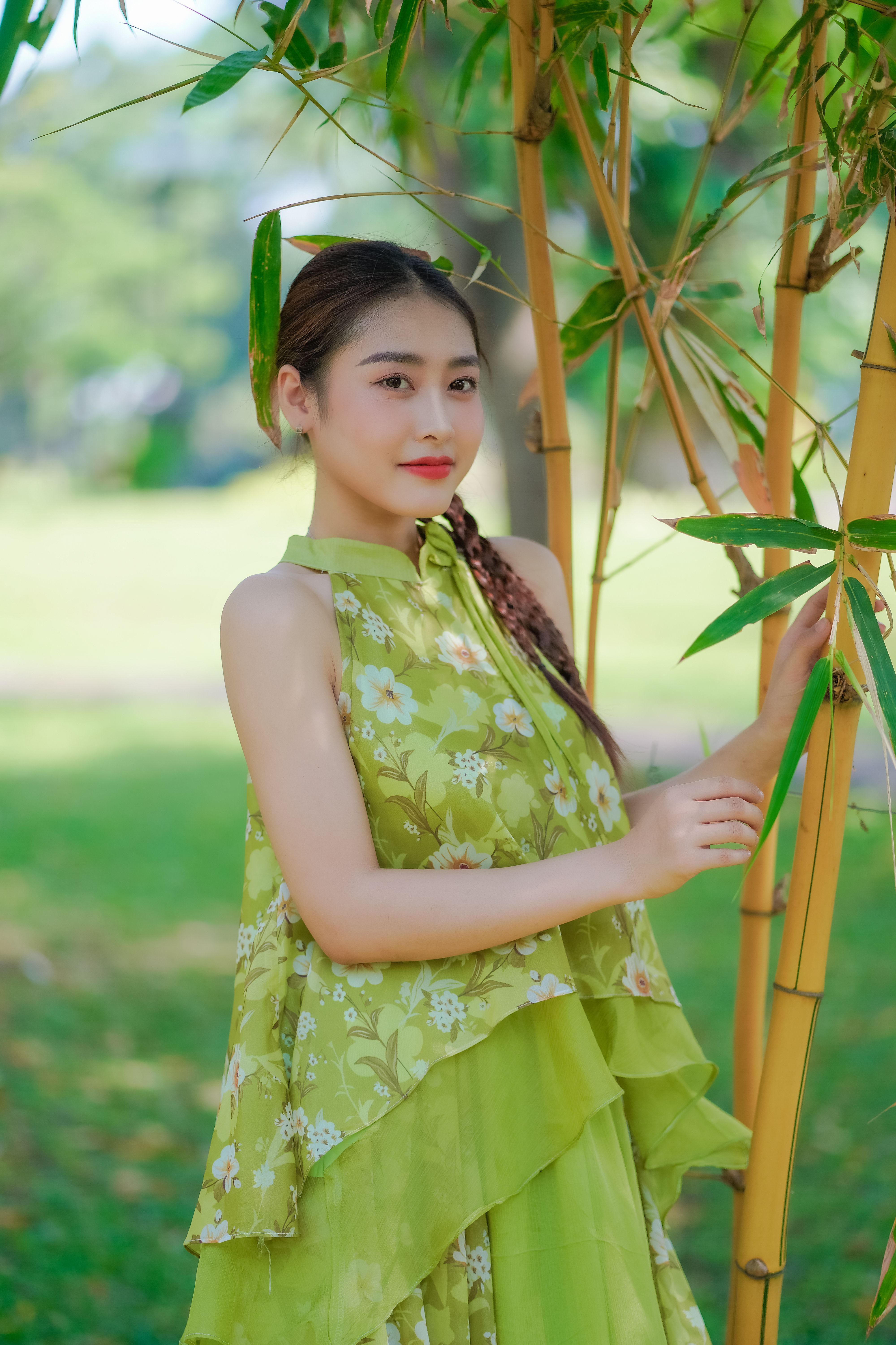 Elegant Asian woman in a green floral dress standing by bamboo outdoors.