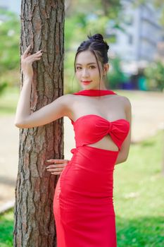 Asian woman in a chic red dress stands elegantly by a tree in a stylish outdoor portrait.
