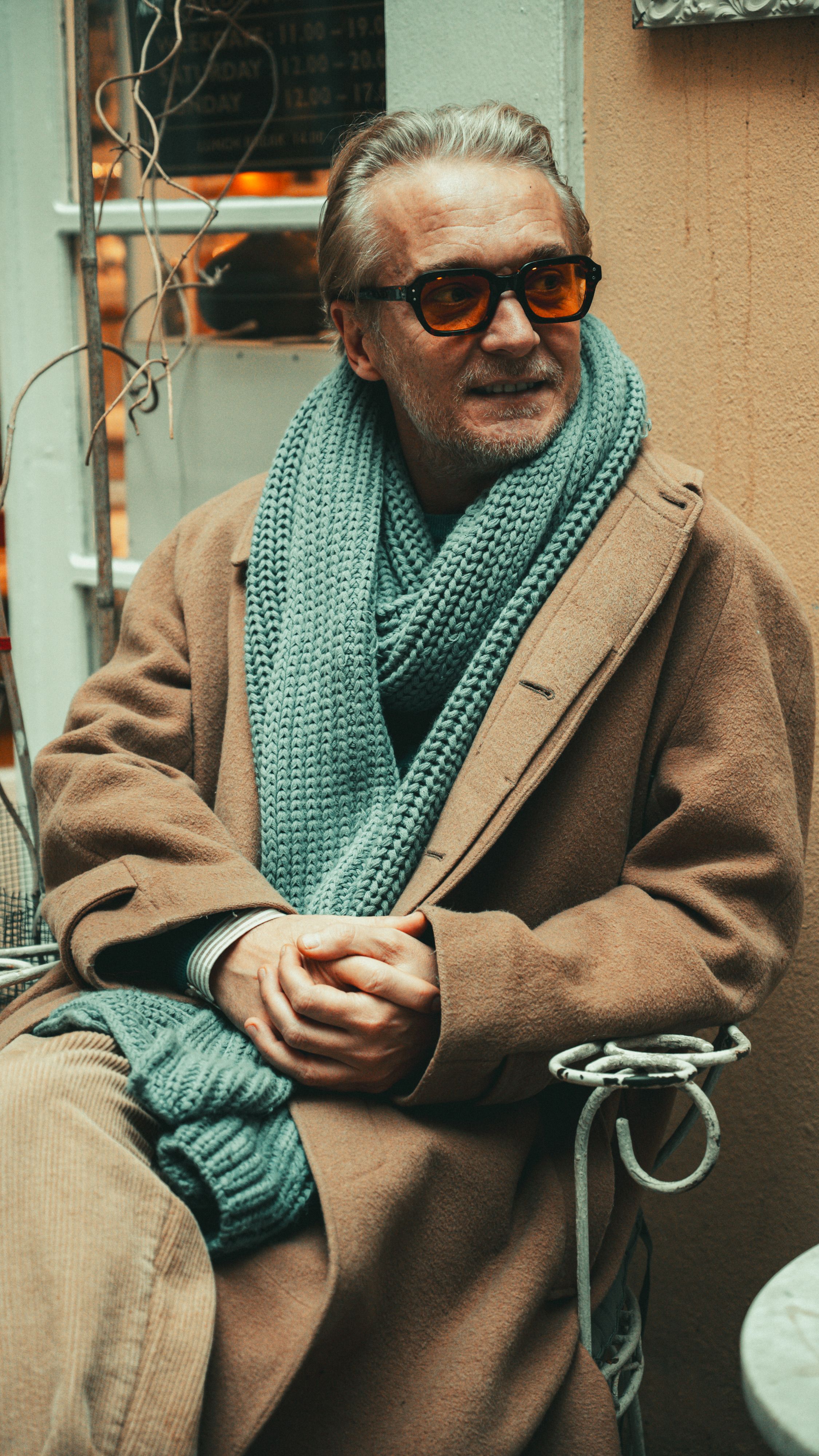 Mature man in brown coat and scarf, sitting elegantly outdoors with sunglasses.