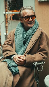 Mature man in brown coat and scarf, sitting elegantly outdoors with sunglasses.