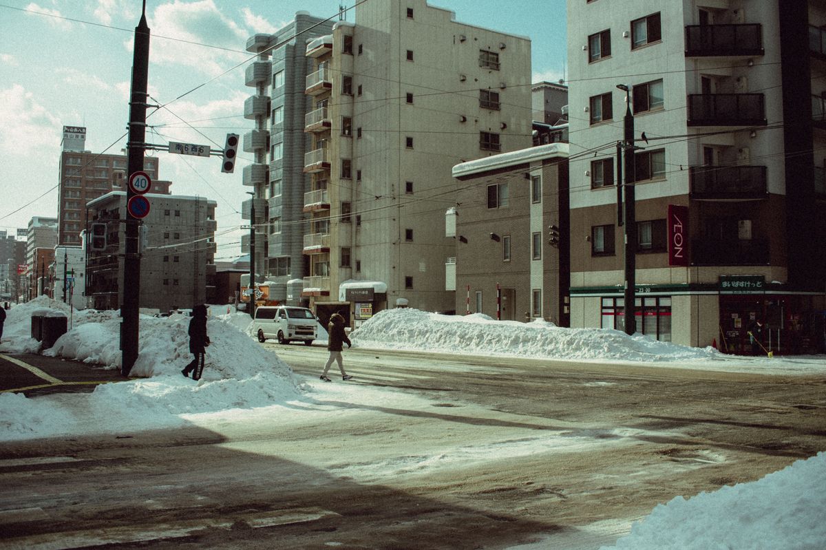 A pedestrian crossing a snow-piled intersection in a Hokkaido city, mid-rise apartment buildings behind and the low winter sun catching the snowbanks.