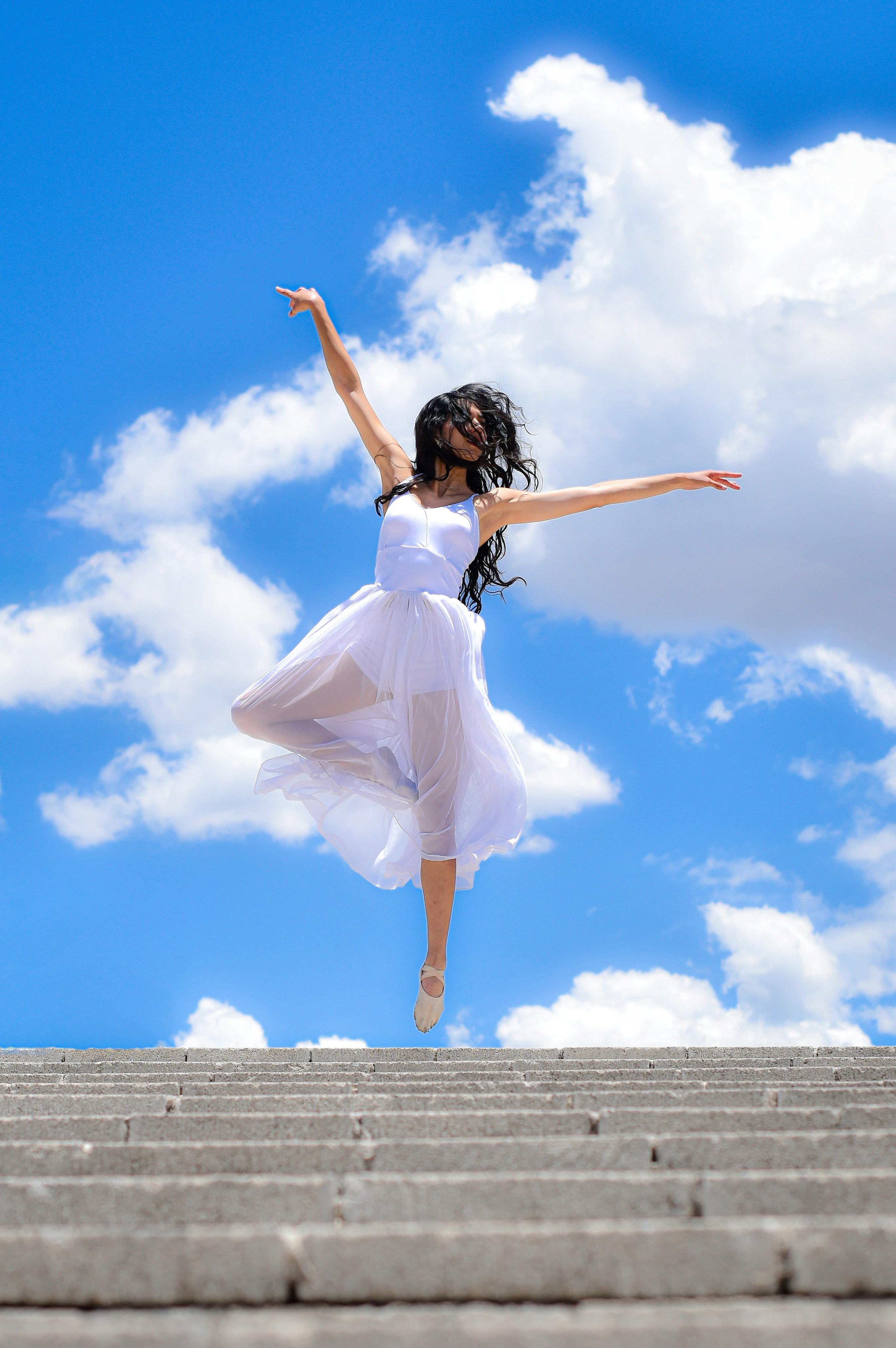 Woman in white dress leaps gracefully against a bright blue sky with fluffy clouds.