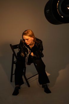 Blonde woman in a stylish black outfit sitting on a chair in a studio setting, dramatic lighting.