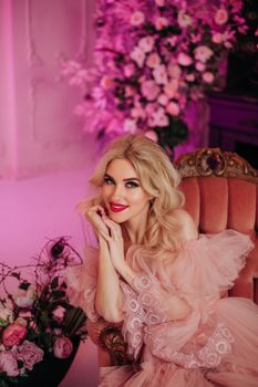 A stylish woman in a pink maxi dress sitting on a vintage armchair surrounded by floral decor.