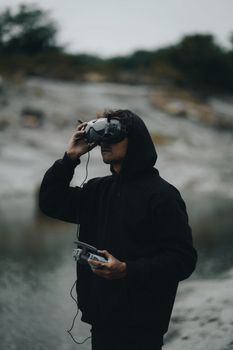 Young man in a hoodie flying a drone using VR goggles, in an outdoor setting.