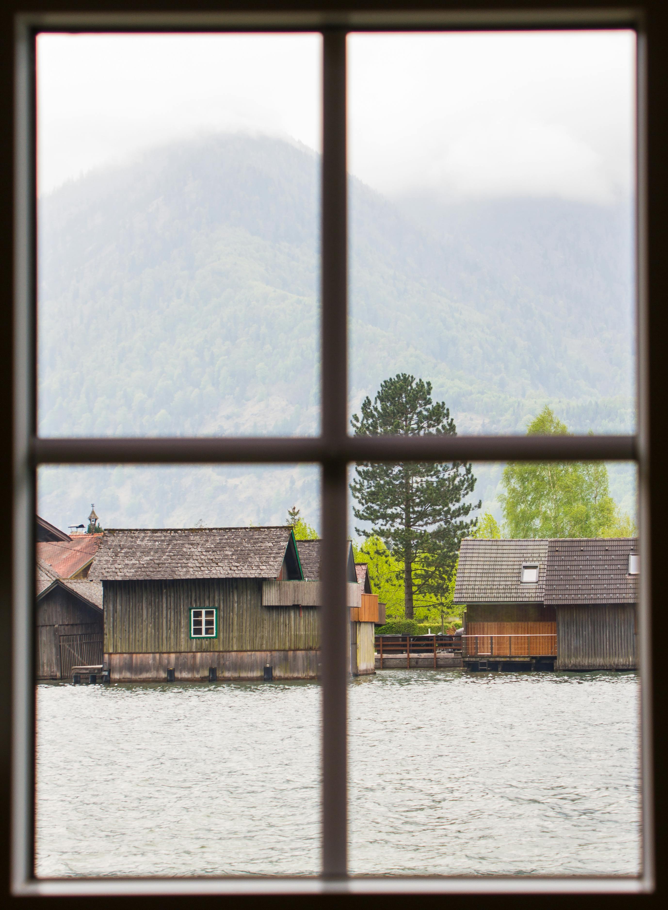 A serene view through a window overlooks lakeside wooden houses with misty mountains in the background.