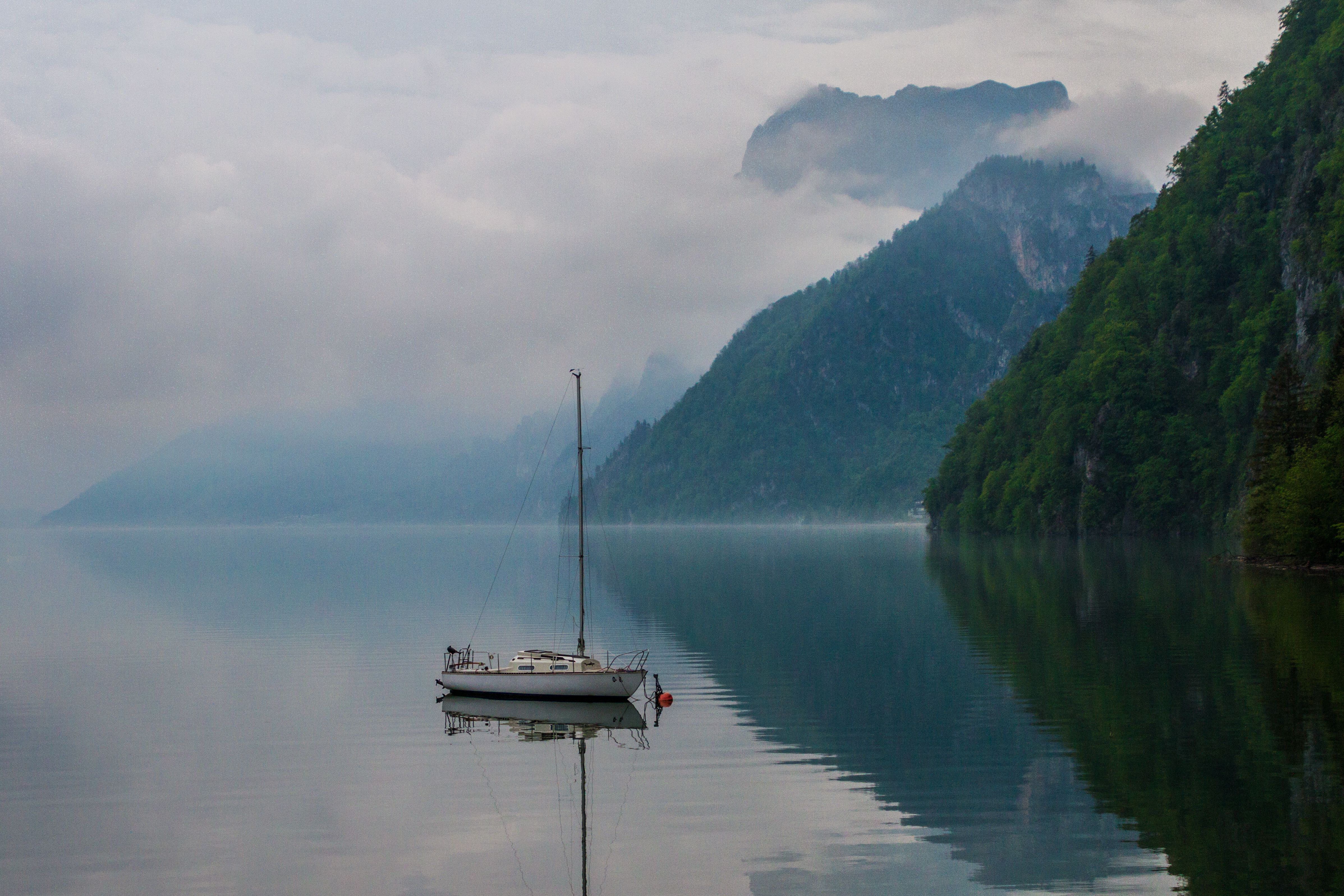 A serene sailboat anchored on a calm, misty lake with mountains shrouded in fog.