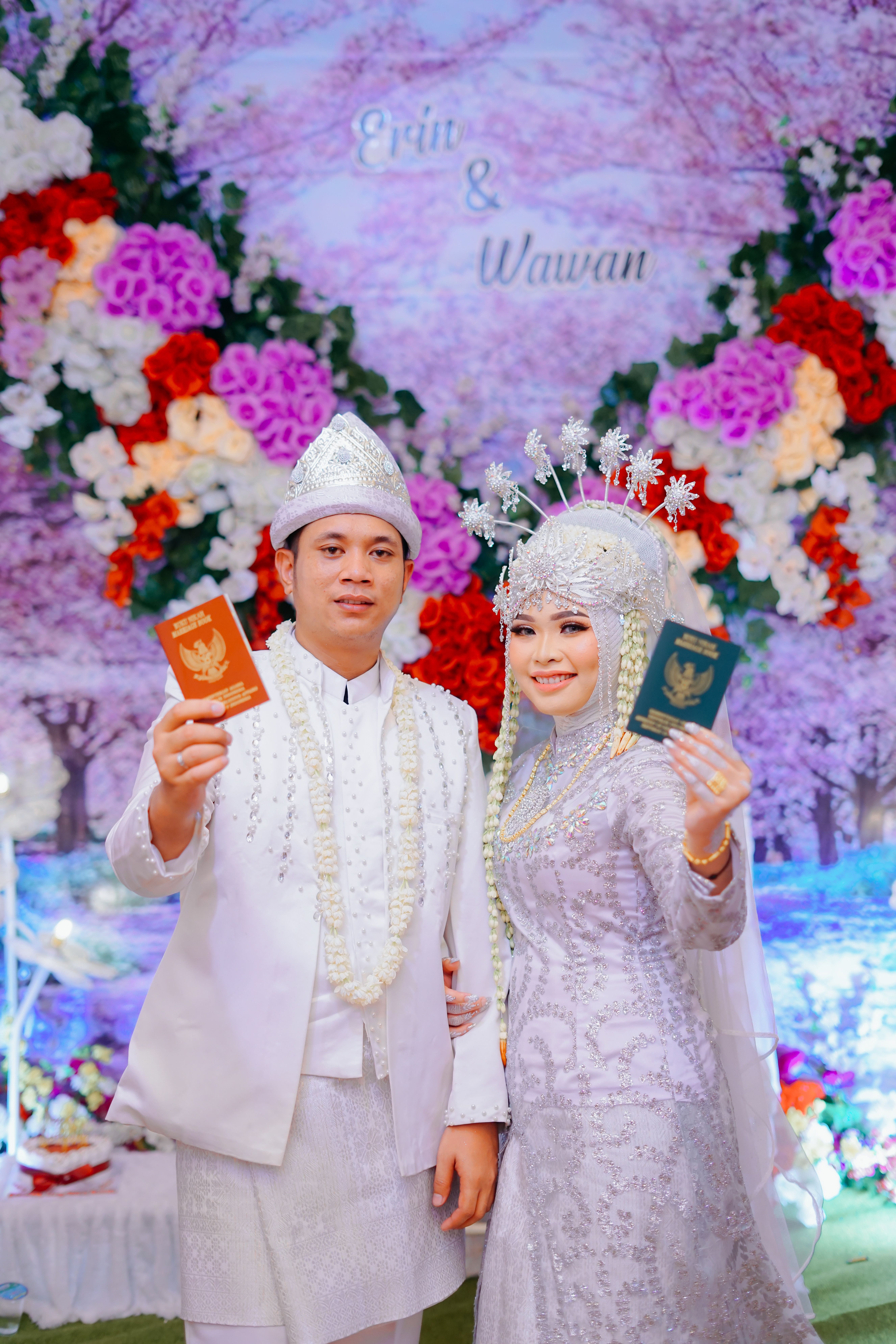 Indonesian couple in traditional attire holding passports at a vibrant wedding celebration.