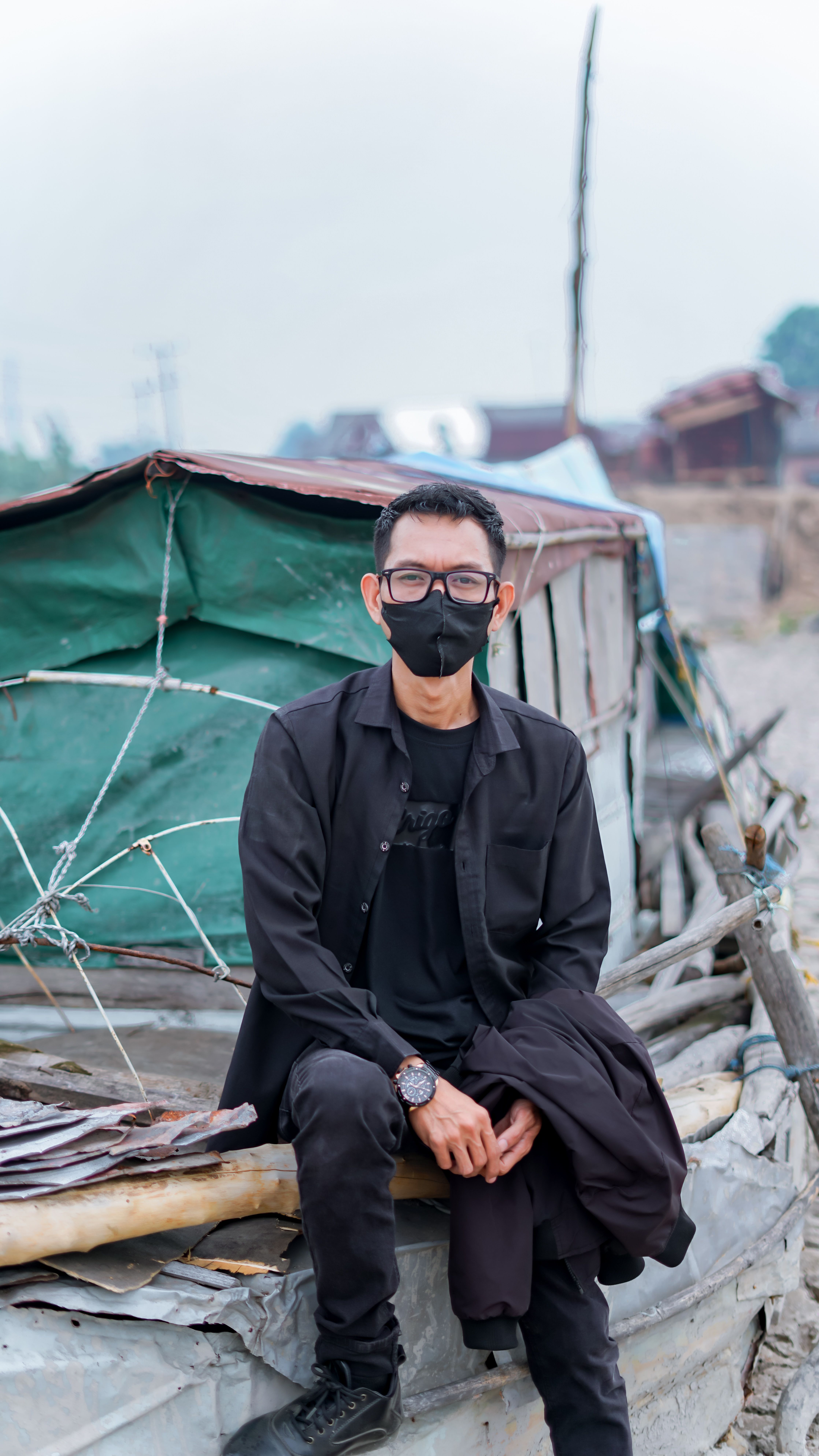 A man in black attire sits on a boat wearing a face mask, outdoors.