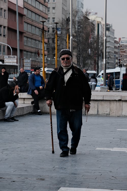 Photo of an Elderly Man with a Walking Stick on a Pavement in City 