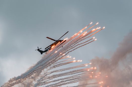 A military helicopter releases flares in the sky, creating a dramatic display against the clouds.