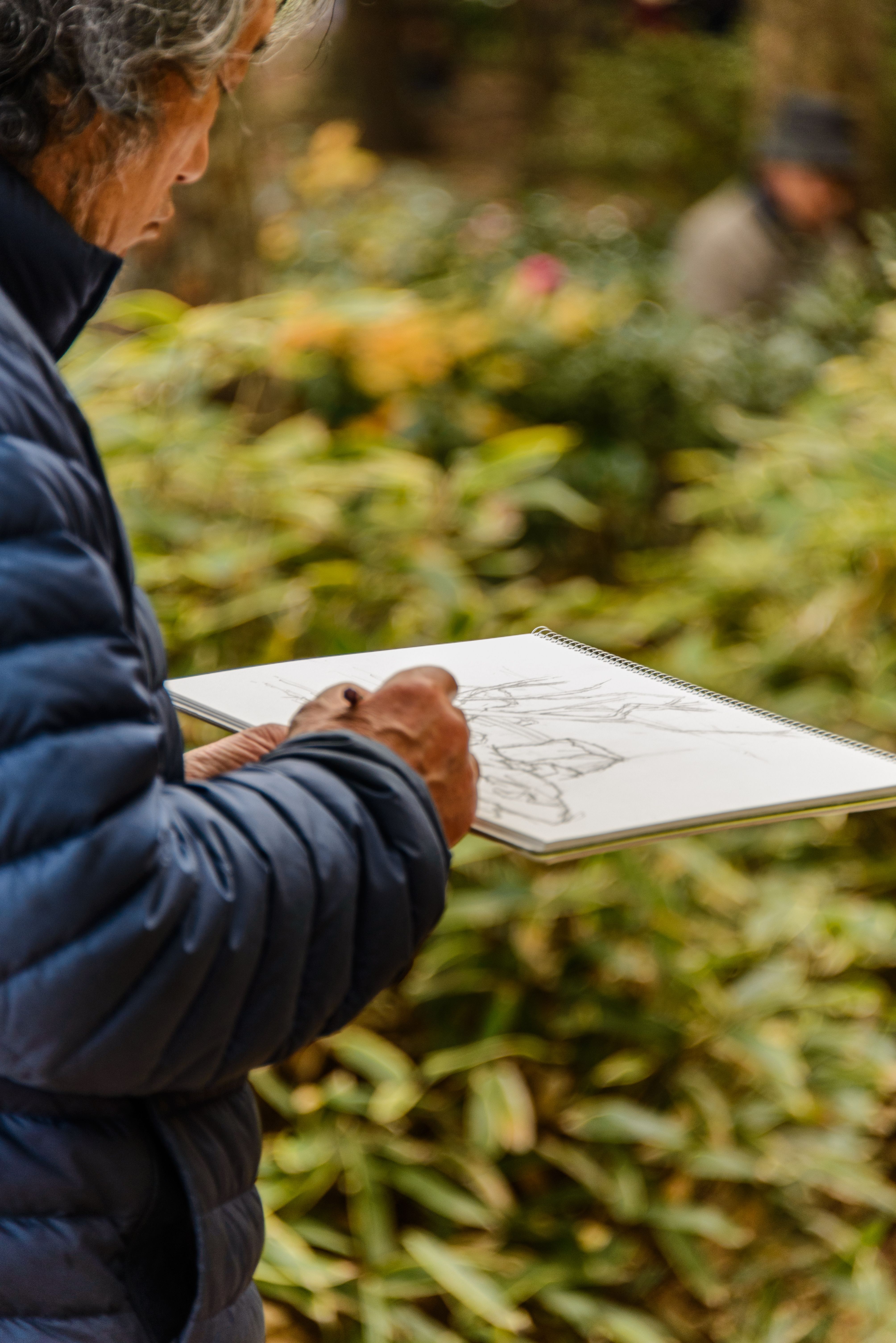An elderly man in a jacket sketching outdoors in a Tokyo park. Focus on creative process.