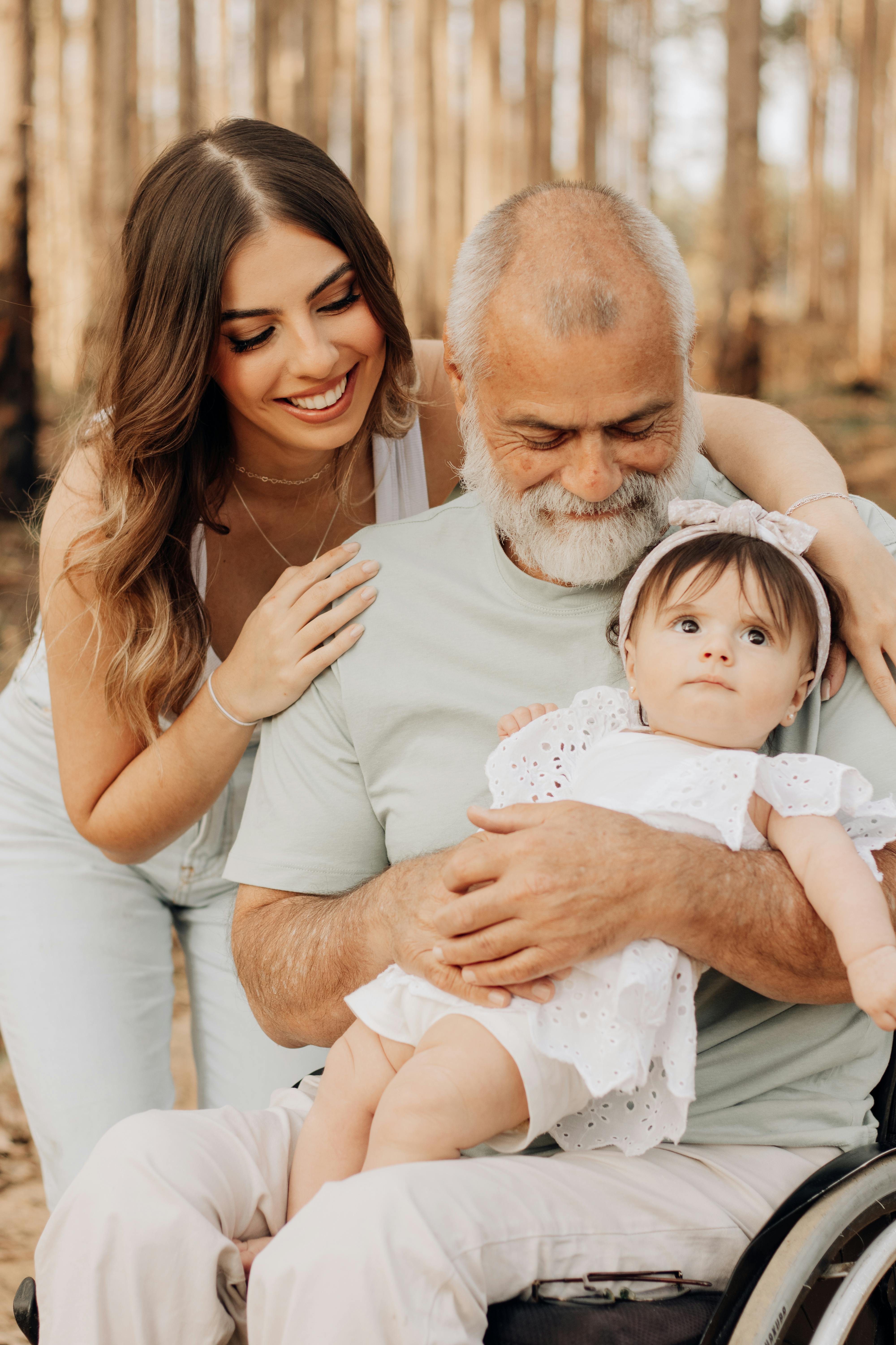 Woman and baby with older man in wheelchair; all are smiling outside in a forest.