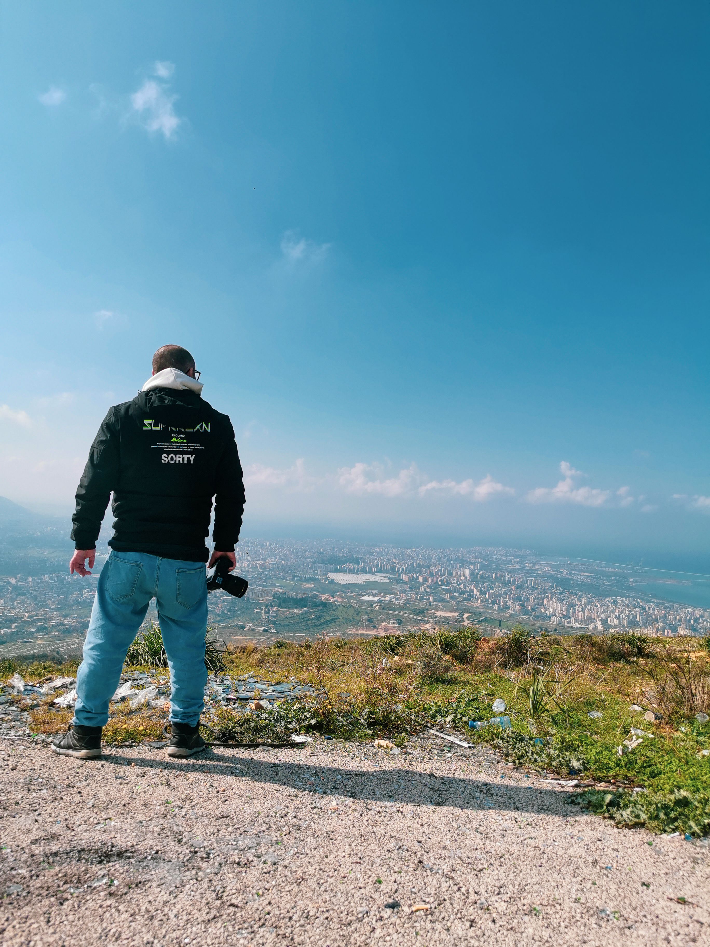 Photographer standing on a hilltop with a camera, overlooking a sprawling cityscape under a blue sky.
