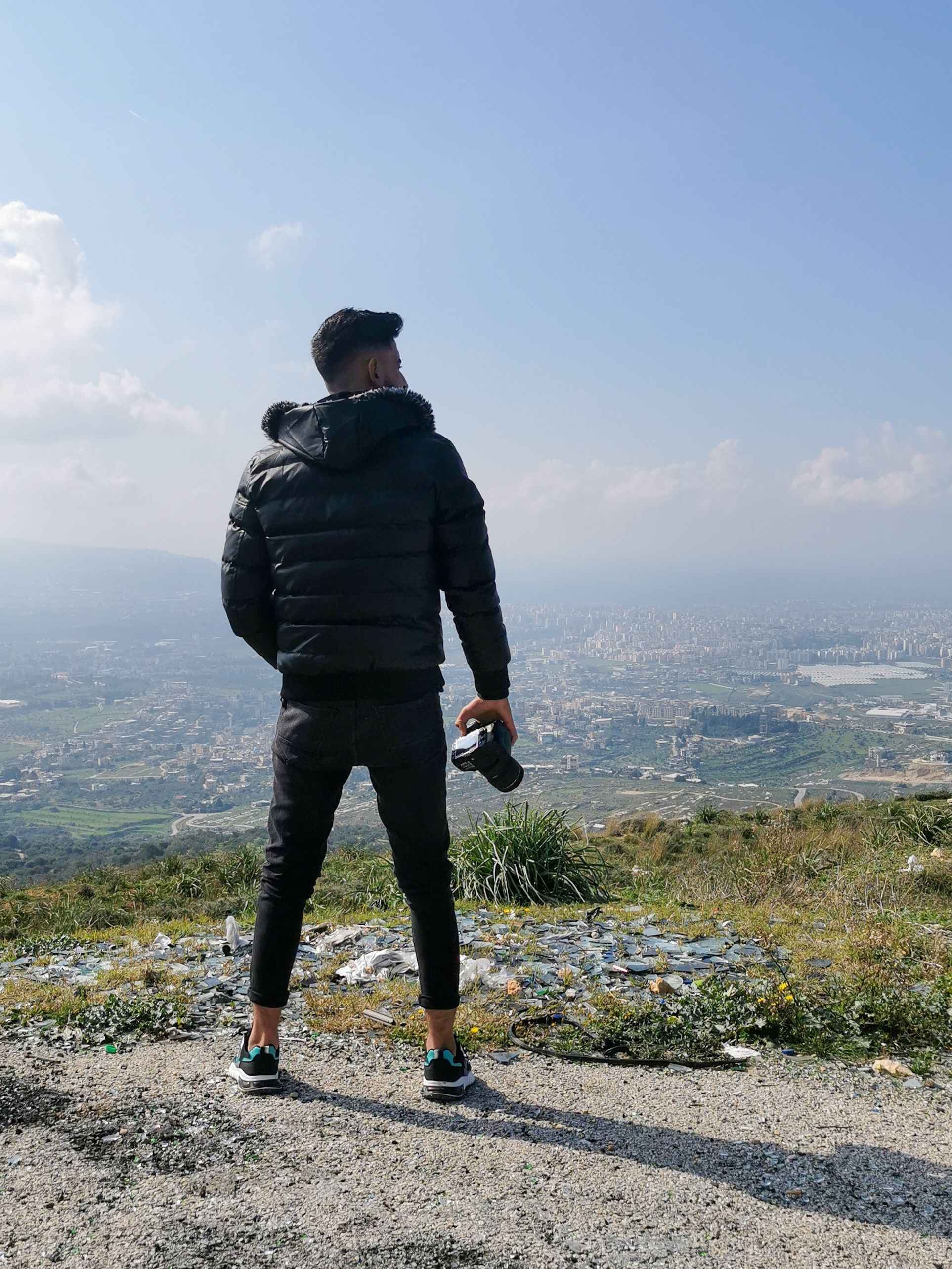 Man in black jacket with camera views city from hillside on a clear day.