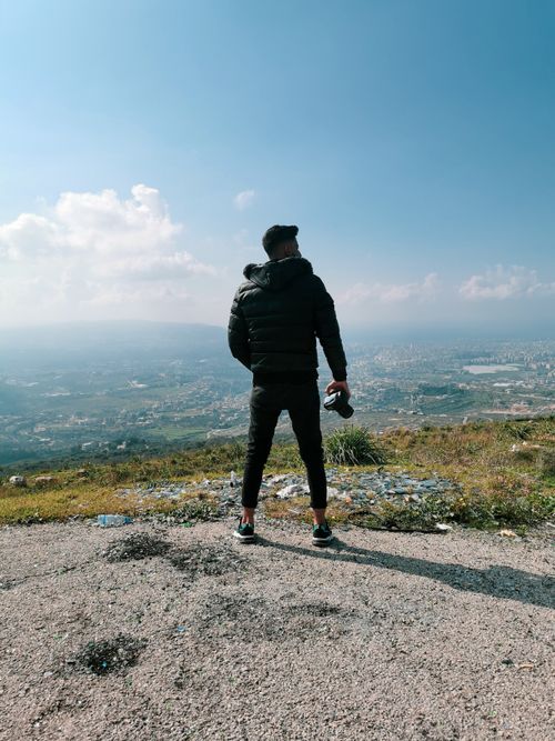 Free A man in a jacket stands on a hilltop, holding a camera, overlooking a vast rural landscape. Stock Photo