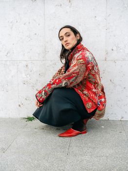A stylish woman poses against a wall in Lisbon, wearing a patterned jacket and red shoes, showcasing urban fashion.