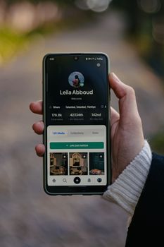 Person holding smartphone displaying user profile, outdoors with bokeh background.