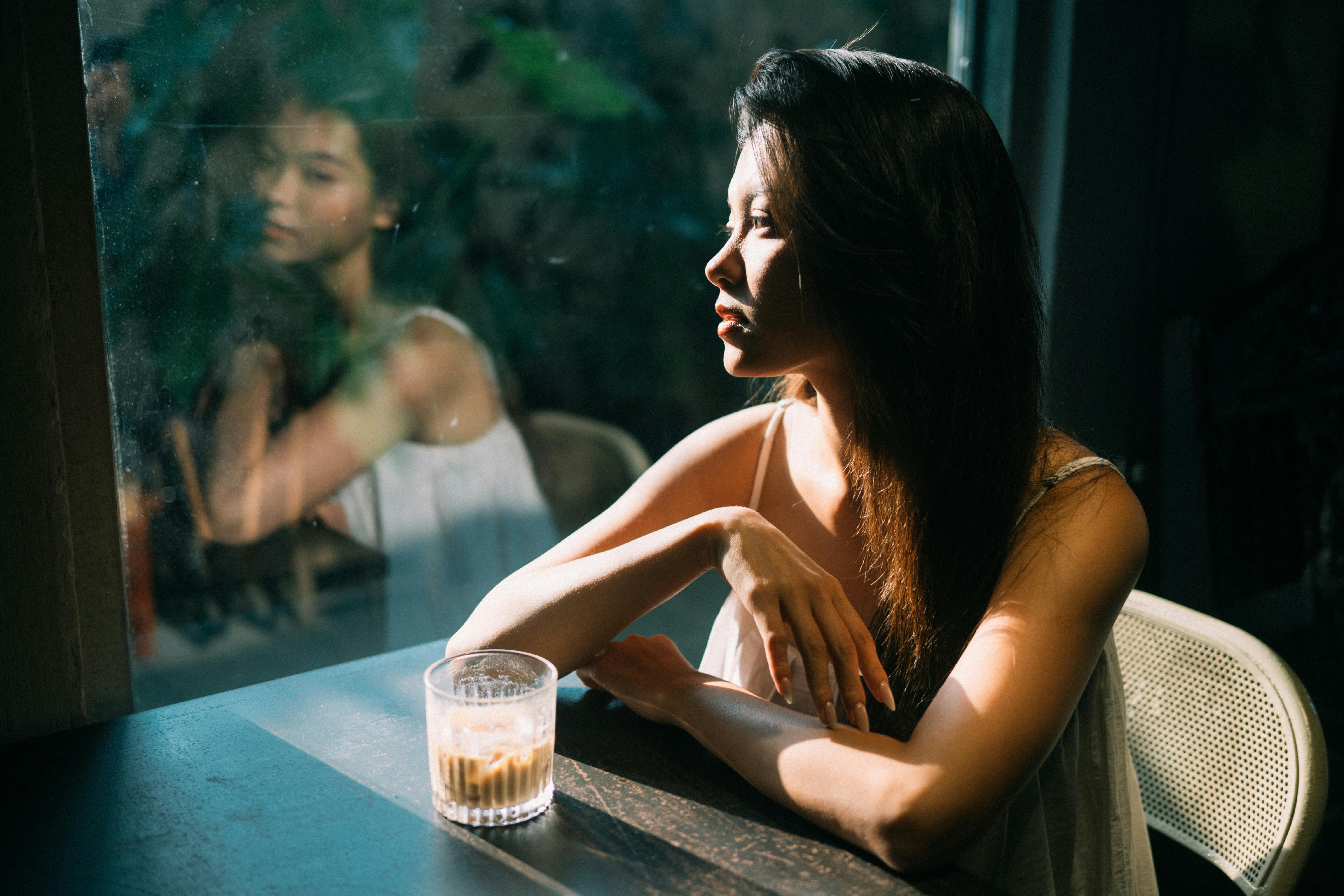 Woman sitting by window with coffee, lost in thought. Soft evening light adds to the contemplative mood.