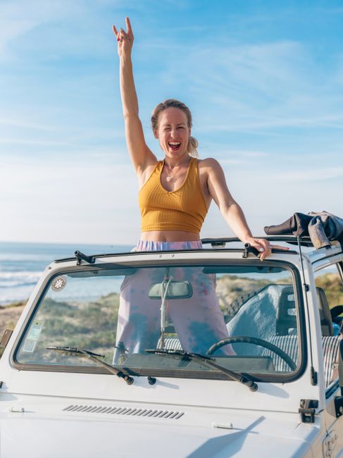 Smiling woman standing in a convertible car near the ocean, celebrating and having fun in Lisbon.