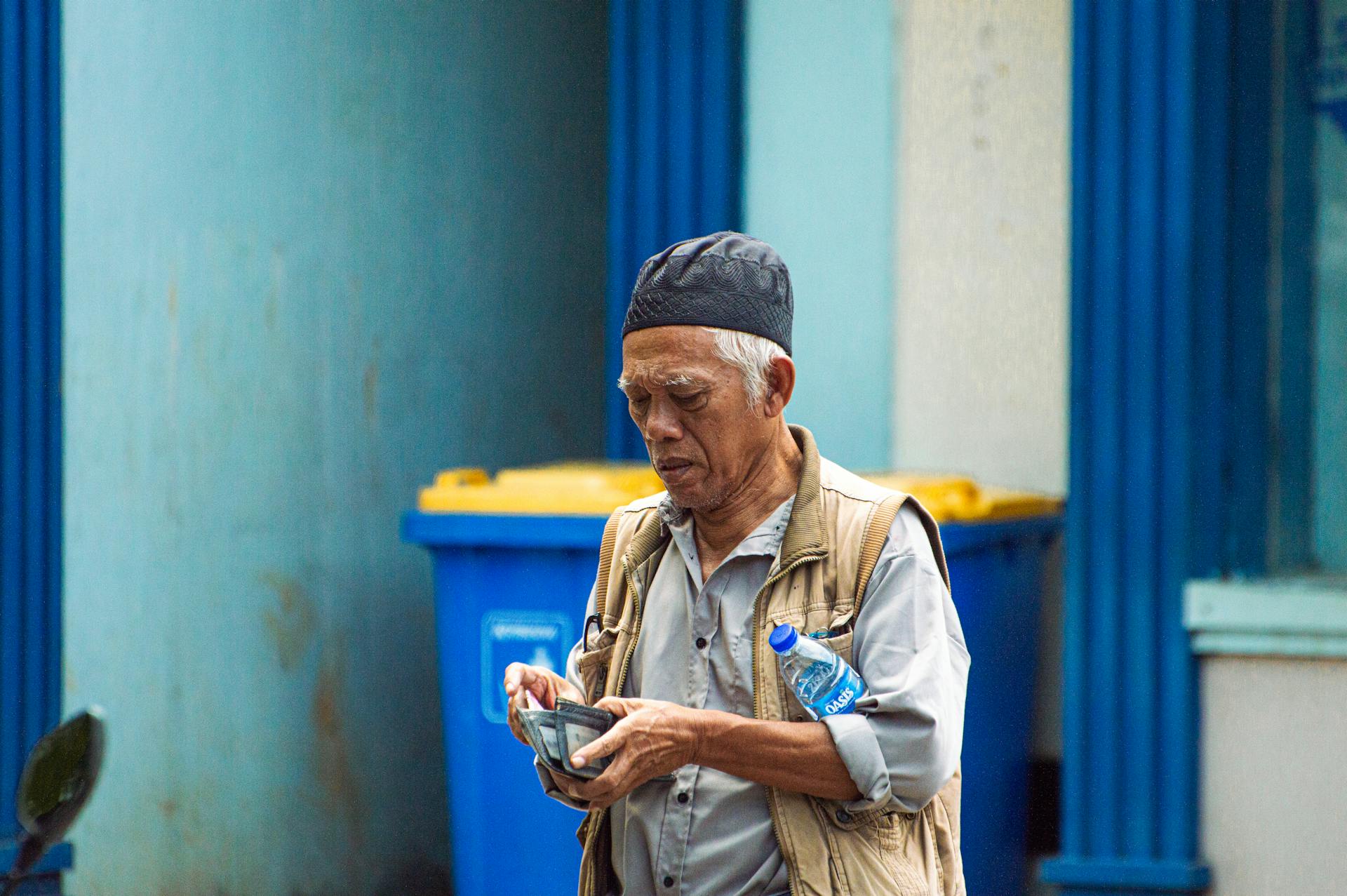 Elderly man wearing a cap counts money while holding a wallet on a city street.