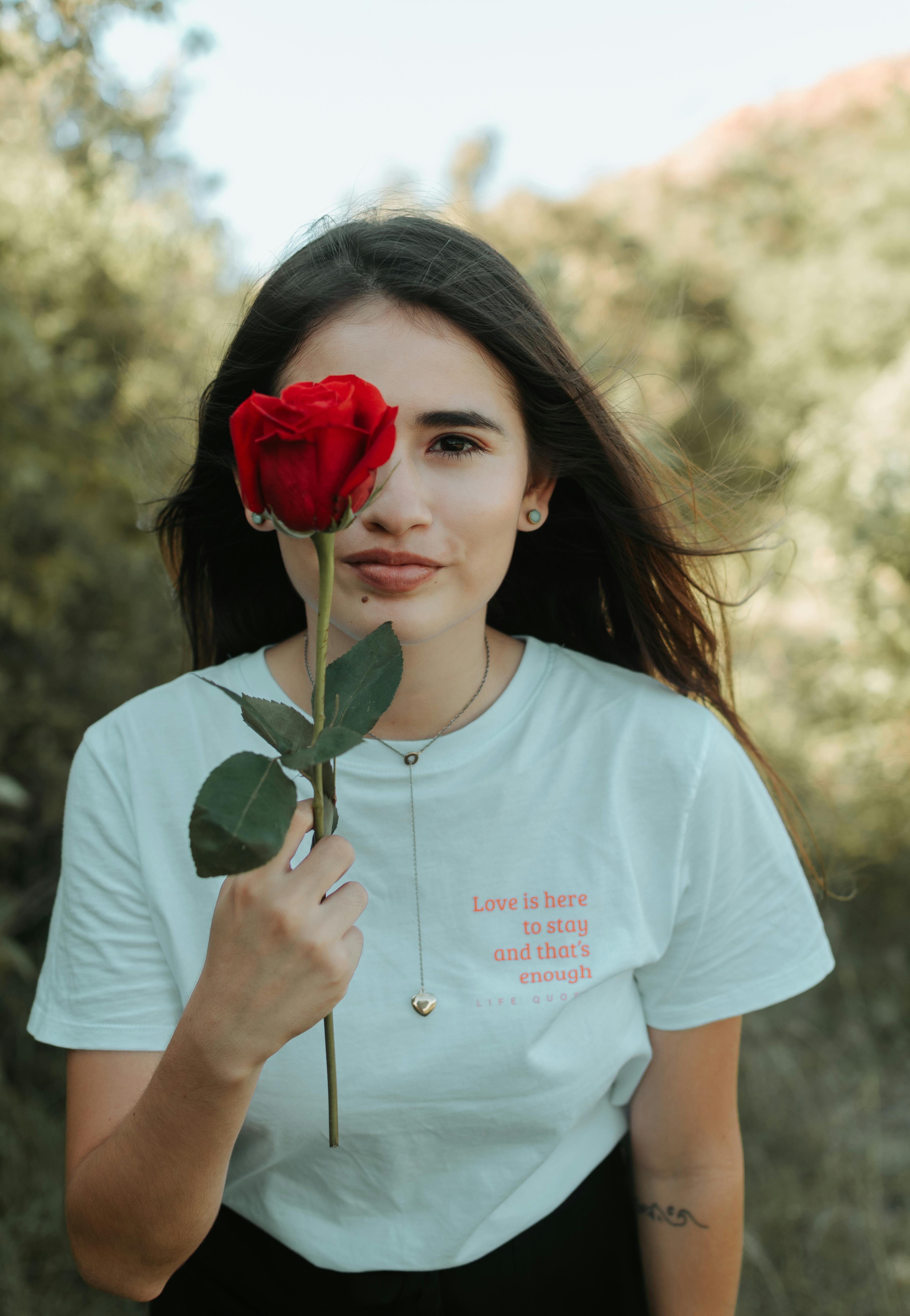 A woman outdoors holds a red rose, showcasing her serene beauty in nature.