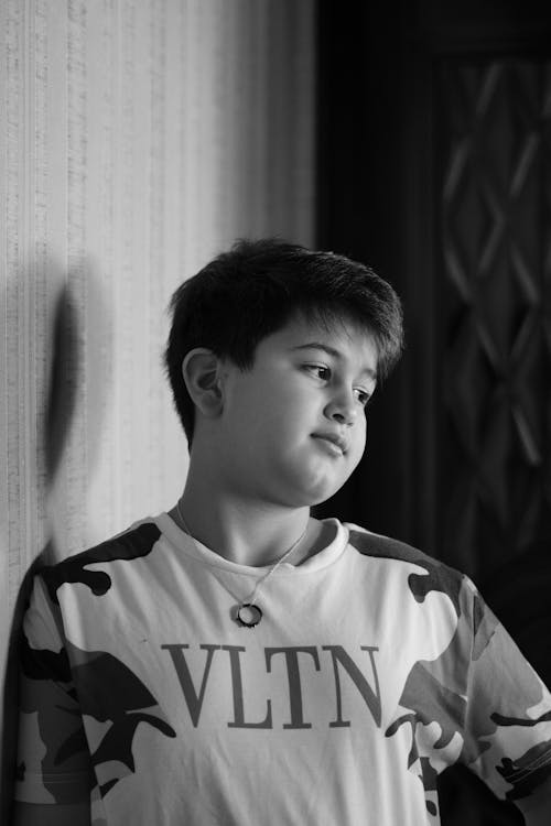 Black and White Photo of a Young Boy Standing against a Wall 