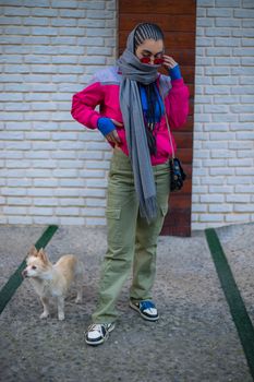 A fashionable woman in vibrant clothing stands with a small dog in an urban environment.