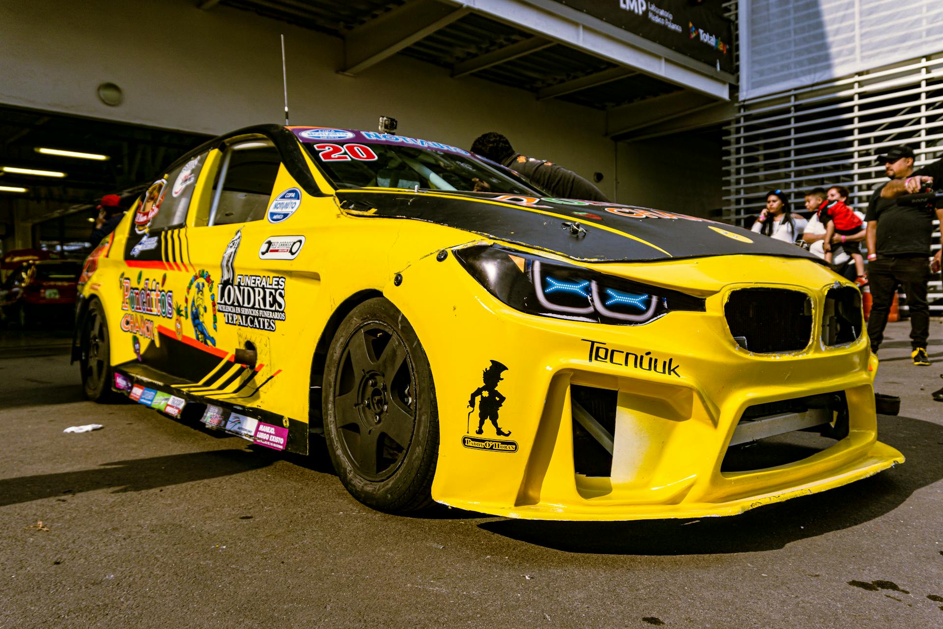 Close-up of a yellow race car with custom decals parked in a garage, surrounded by people.