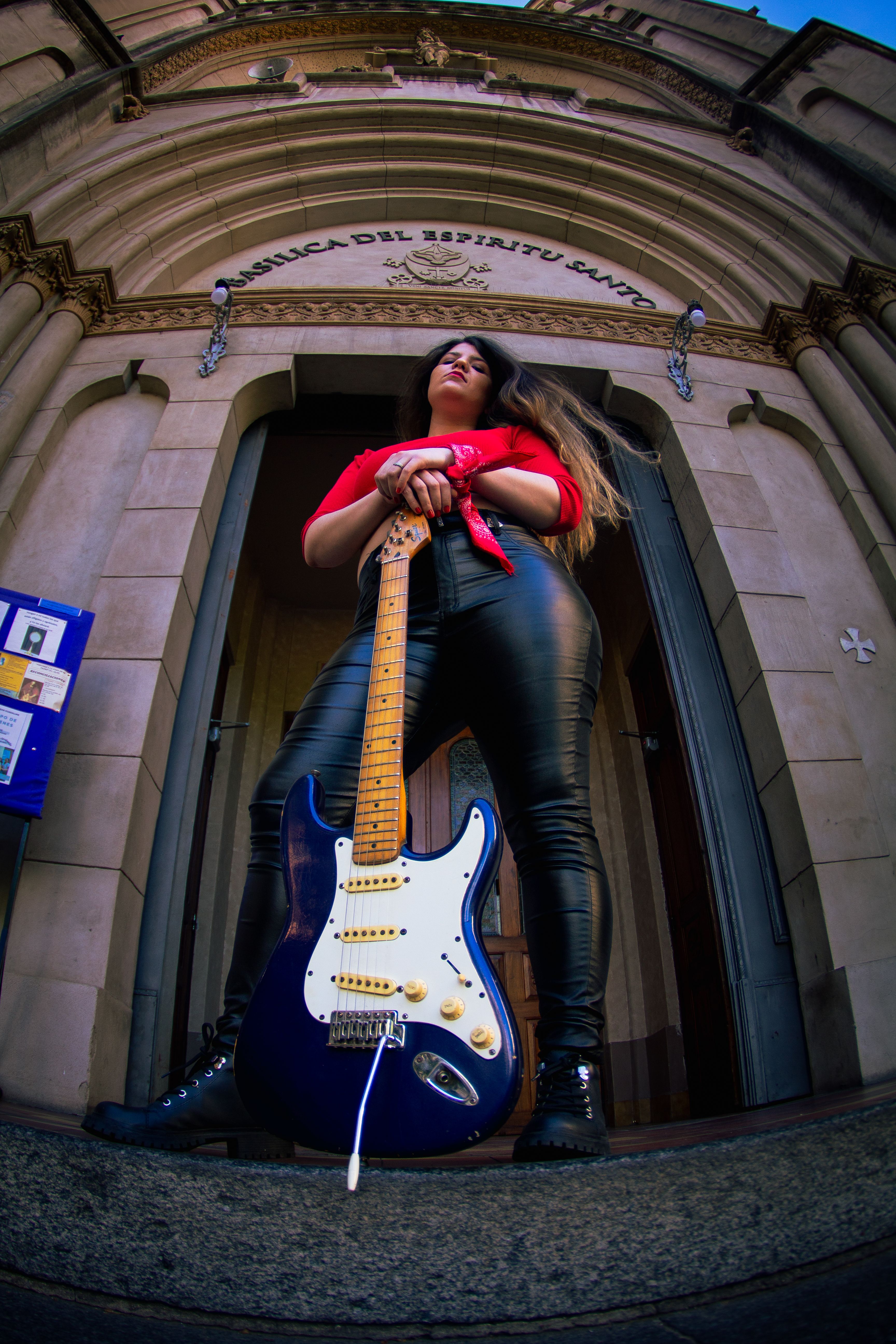 A woman holding an electric guitar stands in front of a grand architectural entrance.