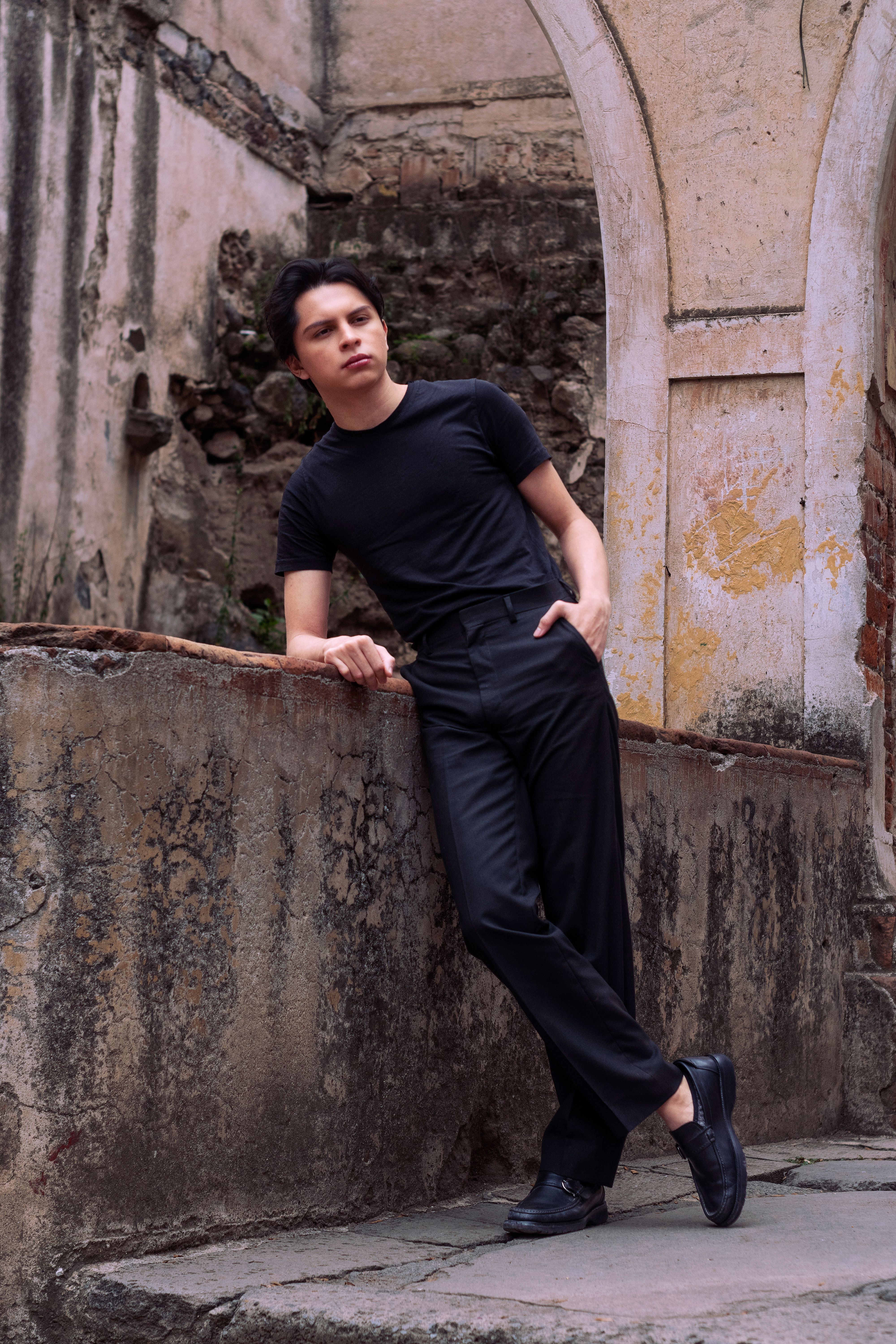 Young man in black attire posing against an old wall in Texcoco, Mexico, showcasing urban fashion.