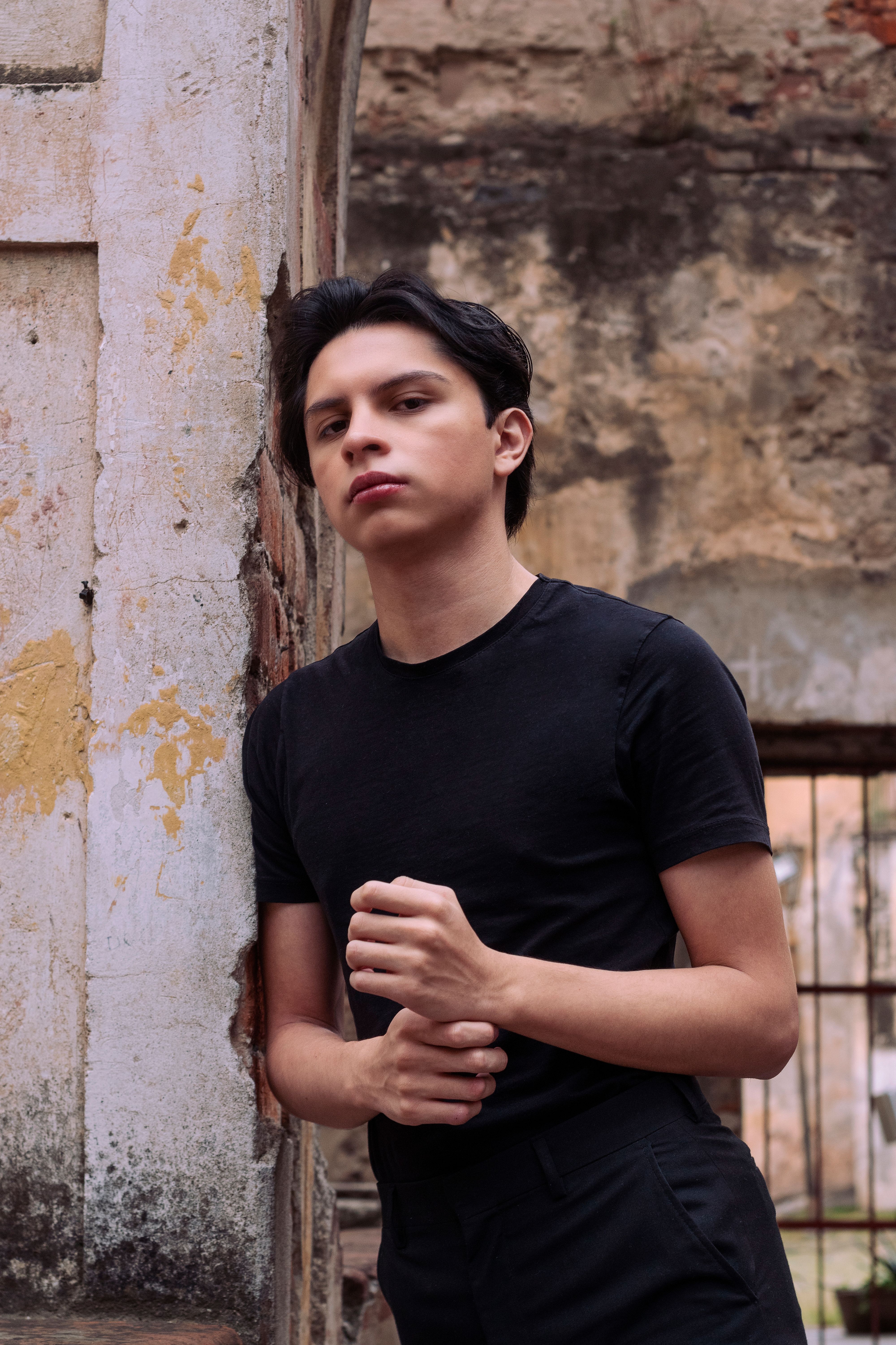 Teenage model in a black t-shirt poses against a textured wall, showcasing urban fashion.