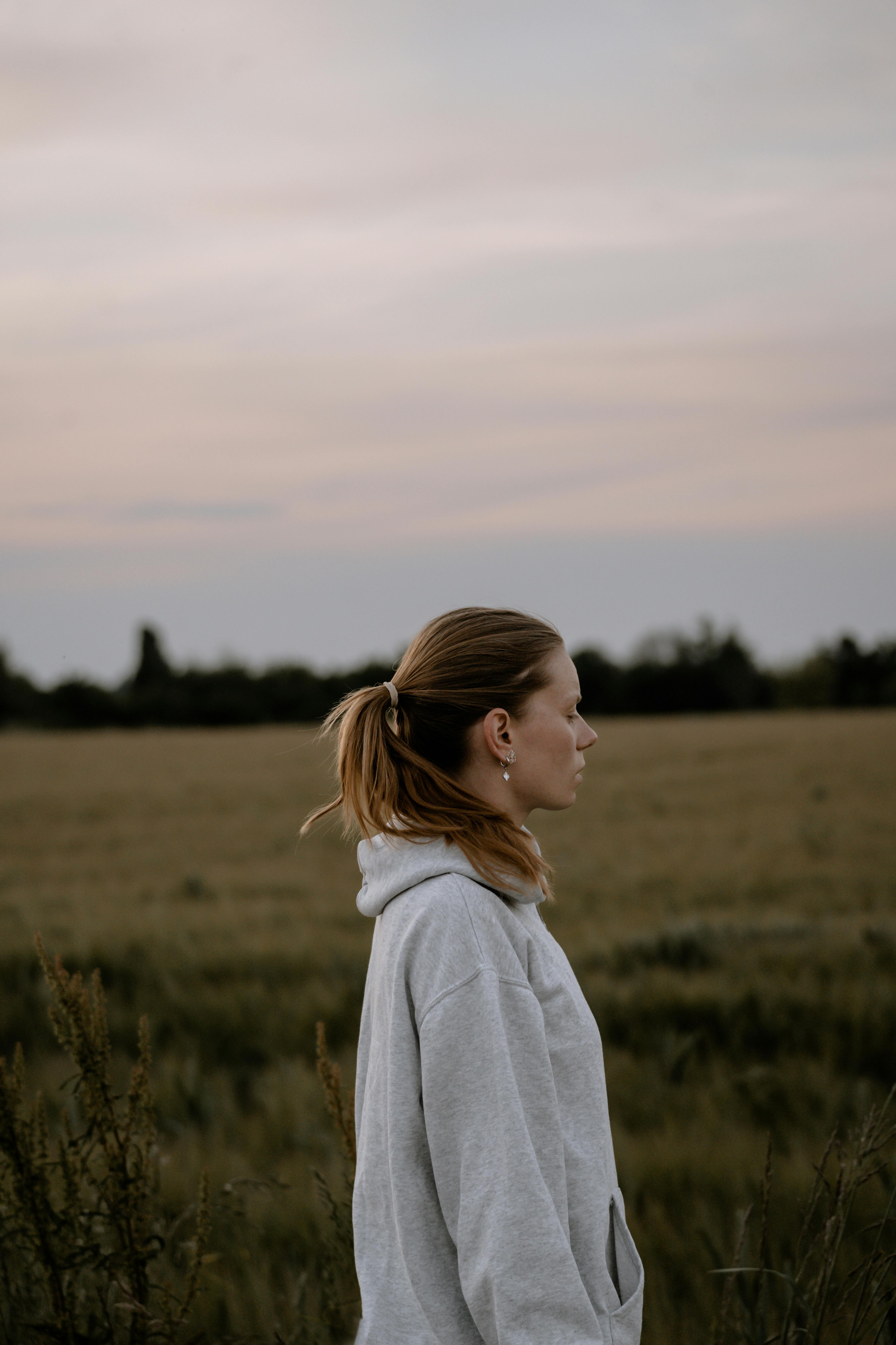 A woman stands in a rural field at sunset wearing a gray hoodie, portraying tranquility and solitude.