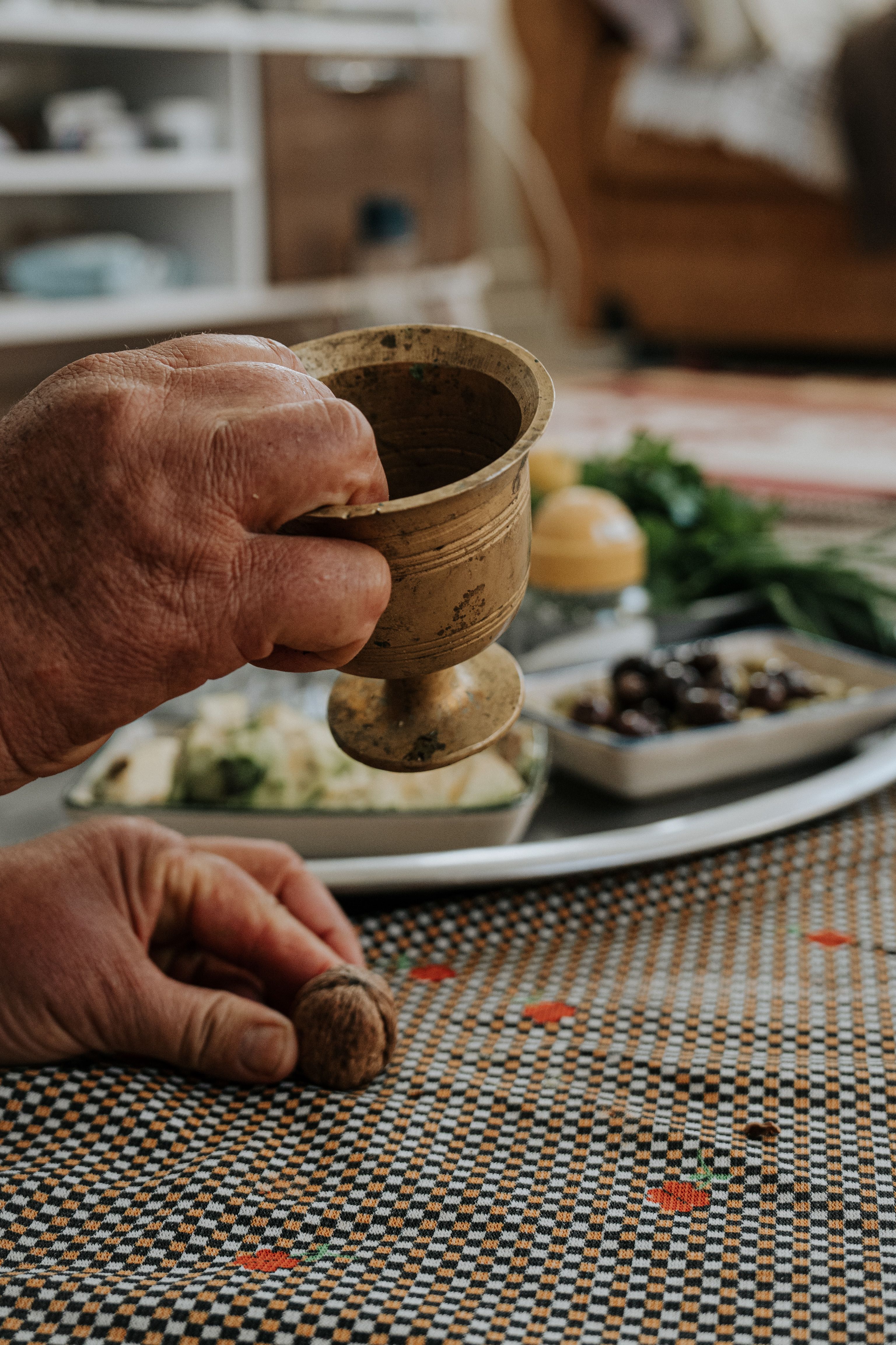 Hands holding a vintage cup and walnut on a patterned table near a food platter indoors.