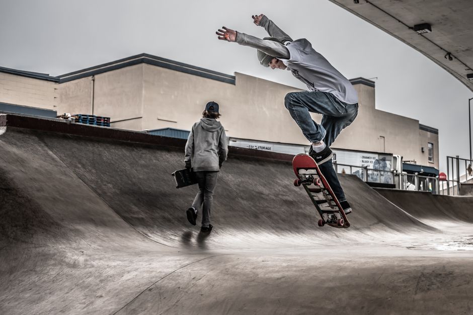 Skateboarder performing an airborne trick at a concrete urban skatepark with gritty, modern architecture.
