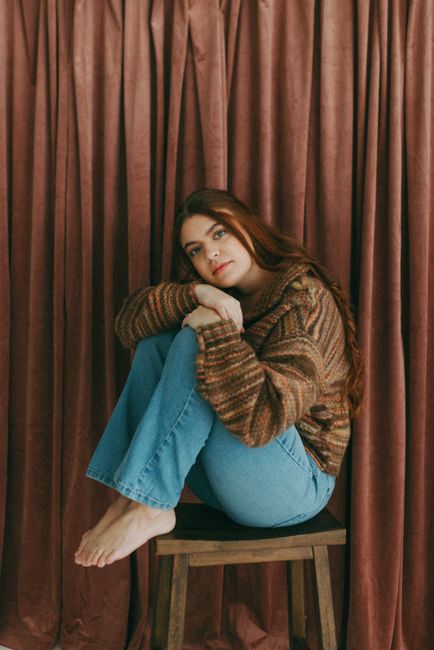 A woman in jeans and a cozy sweater sits on a chair indoors against a fabric backdrop.