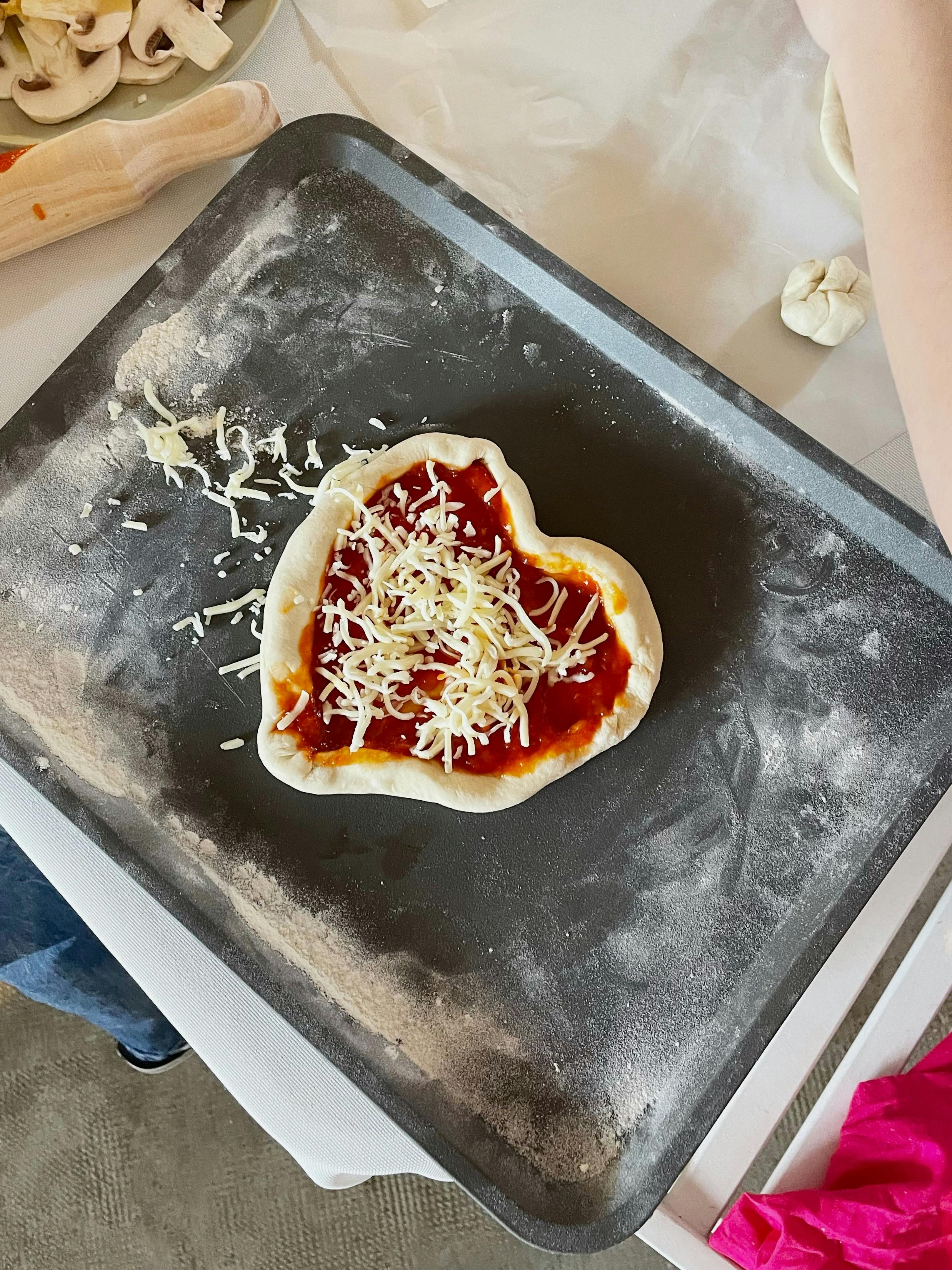 Top view of heart-shaped pizza dough with sauce and cheese on a baking tray, ready to bake