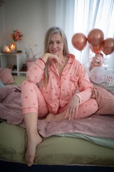 Blonde woman in pink pajamas smiling while sitting barefoot in a cozy room with balloons.