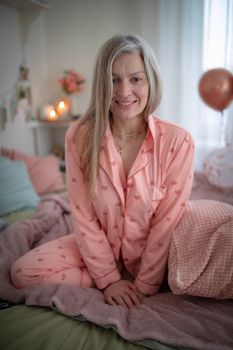 Blonde woman in pink pajamas sitting on a cozy bed with a cheerful smile, surrounded by soft lighting.