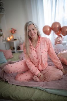 Woman in cozy heart-print pajamas sitting on a bed surrounded by balloons and soft lighting.