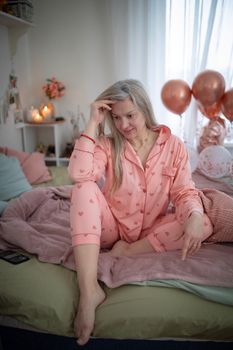 Blonde woman in heart-patterned pajamas relaxing on bed with pink and rose gold decor.