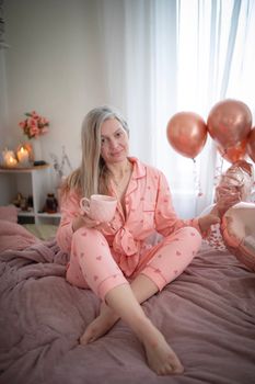 Woman in pajama holding coffee cup surrounded by balloons on bed.