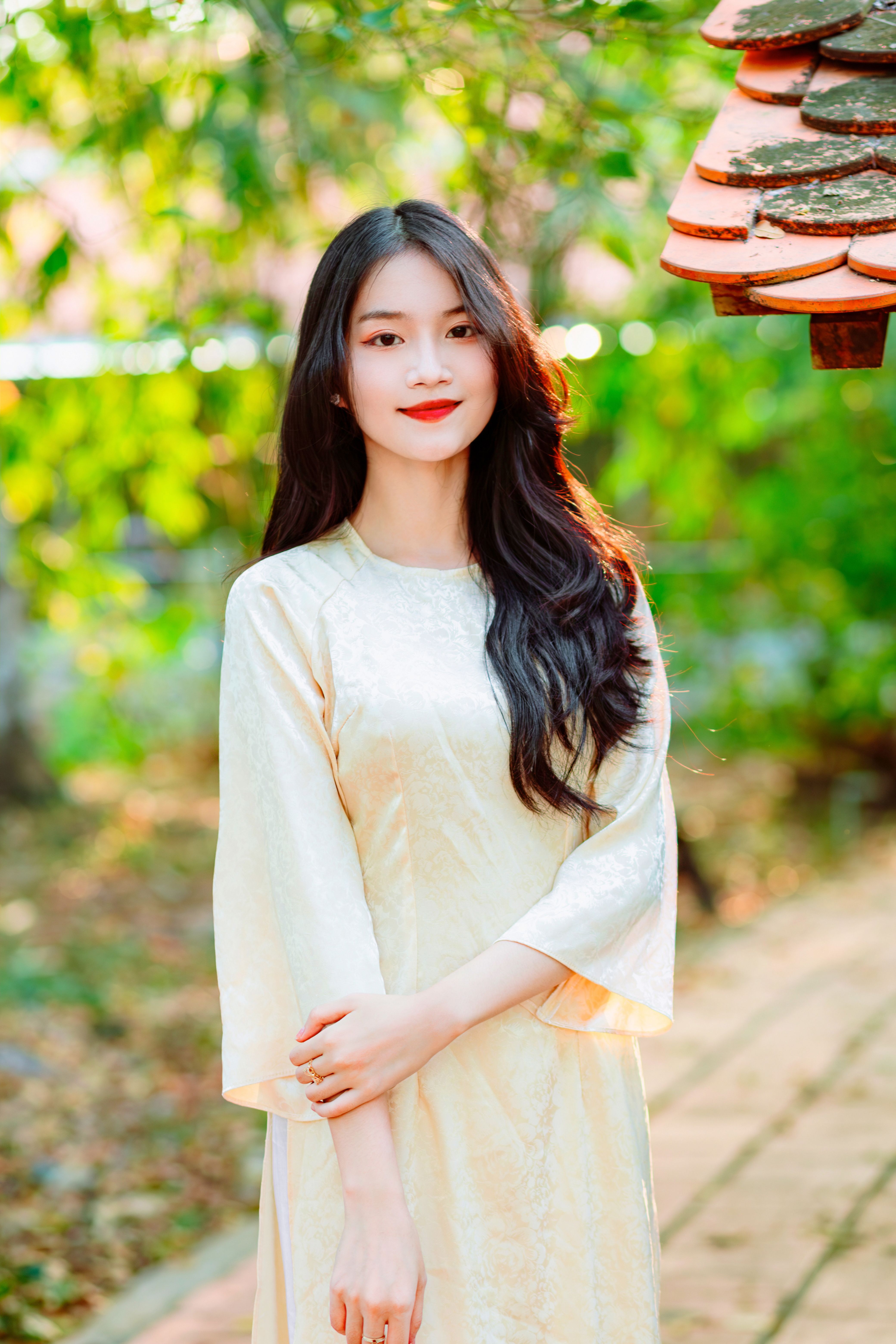 Beautiful woman in cream dress posing gracefully outdoors, surrounded by vibrant greenery.