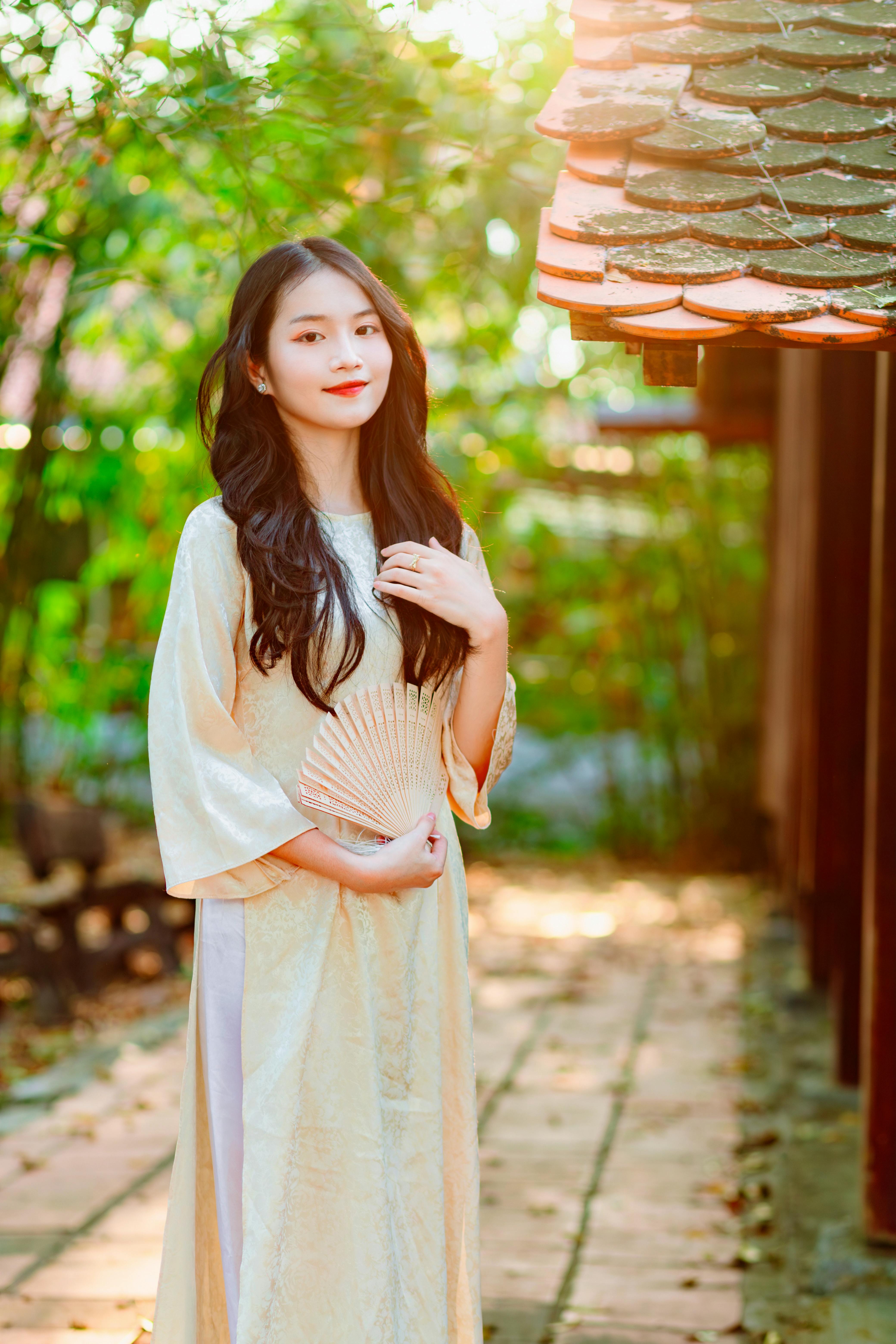 Elegant woman in a traditional white dress holds a fan, standing outdoors in sunlight.