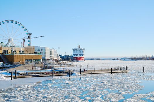 A scenic view of Helsinki's frozen harbor featuring a ferris wheel and a stationary ship.