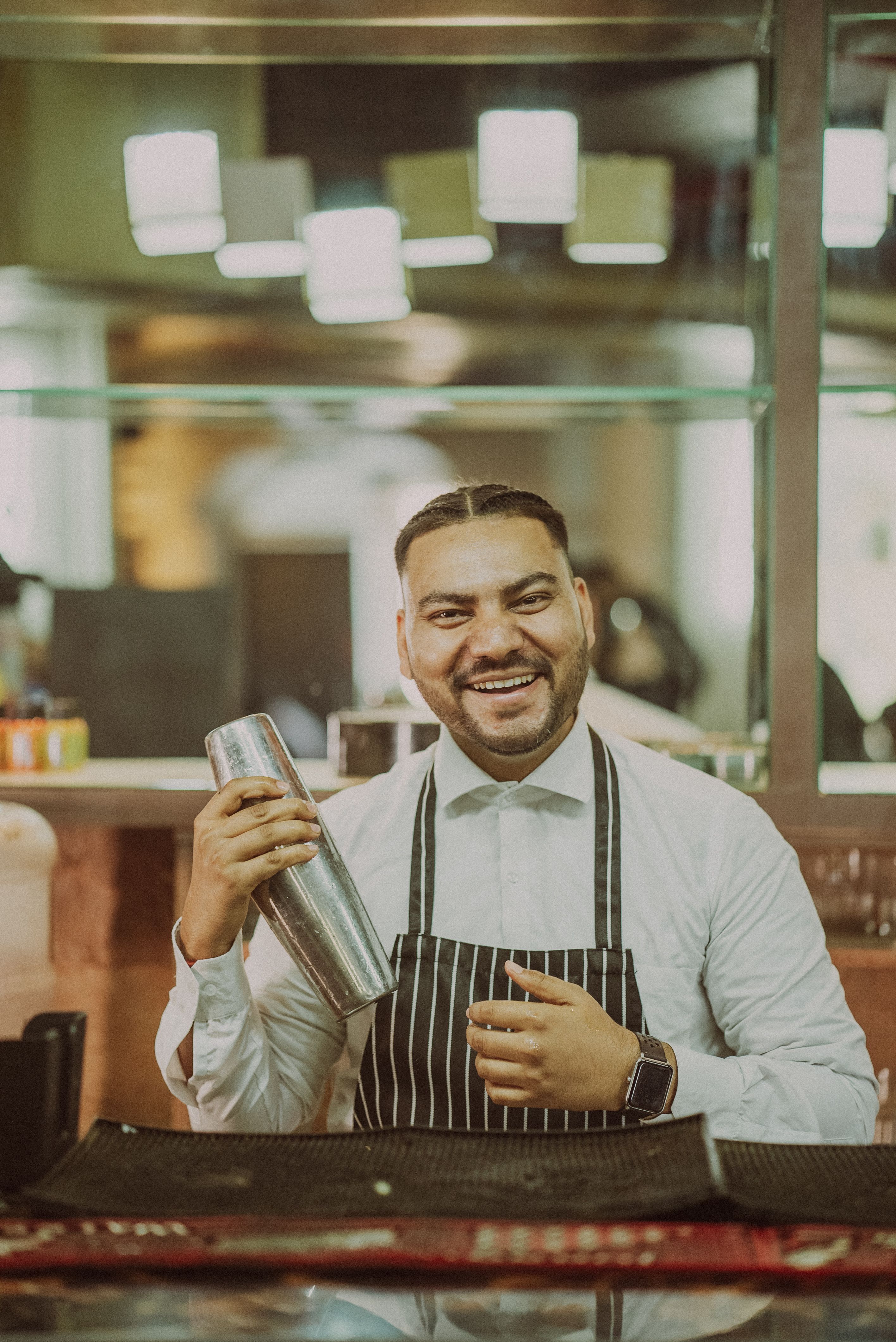 A happy barman in New Delhi mixing cocktails with a shaker, wearing a striped apron.
